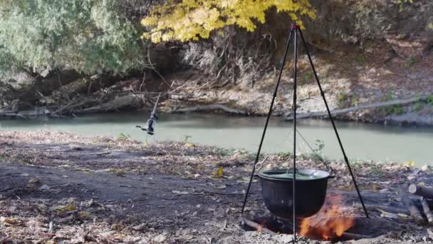 回転棒の近くで木を燃やして蓋をしたカルドロン 川岸で魚スープを調理する — ストック動画