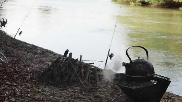 石炭をめぐって沸騰するティーポット 釣り棒と川の背景に対して — ストック動画