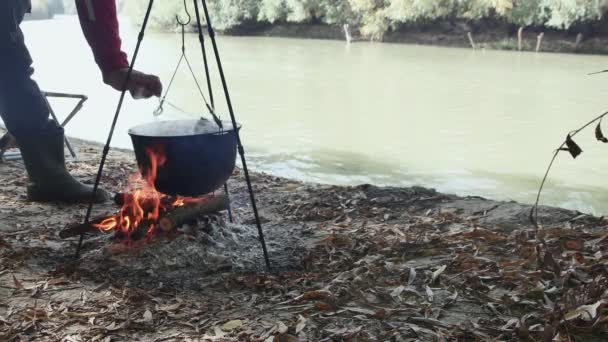 川岸の男が しごで魚のスープをかき混ぜ 自然の中の火でカルドロンで魚のスープを調理する — ストック動画