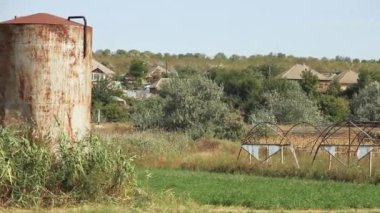 Terk edilmiş bir seranın yanında eski paslı bir Sovyet su tankı kırsal evlerin arka planına karşı.
