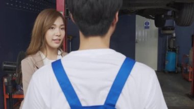 Young man mechanic talking with customer for examining trouble and repairing engine of car in the garage, vehicle of asian woman with problem and maintenance, advice of technician about automobile.