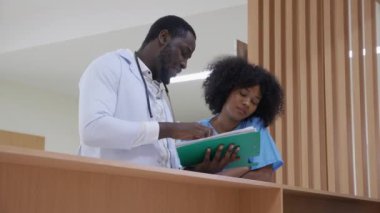 Young African American doctor specialist talking and looking document about patient with nurse or assistant for diagnostic together at corridor in the hospital, medical and insurance concept.