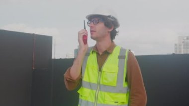 Young caucasian man using radio conversation and inspecting while building and tower all around, engineer or architect examining and maintenance for development, industrial and communication concept.