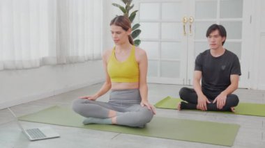 Genç kadın ve Asyalı erkek, evde oturup kaslarını esneterek yoga yapıyorlar. Çift antrenmanı egzersizleri yaparken dizüstü bilgisayar, spor konsepti izliyorlar..