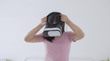 VR kulaklık takan genç Asyalı bir kadın evdeki yatak odasındaki yatakta heyecanla oyun oynuyor. Sanal gerçeklik ya da metaz yeniliklerini kullanarak 3 boyutlu, yaşam tarzları ve teknoloji kavramını kullanıyor..
