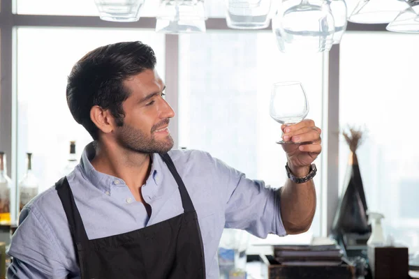 Young caucasian man is bartender holding and looking clean of glass with professional at counter, portrait handsome male preparation glass or wineglass, barman standing with confident at restaurant.