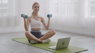 Beautiful young woman sitting on mat and holding dumbbell while using laptop computer for practicing workout in the room at home, female exercise and bodyweight training for health, sport concept.