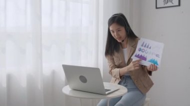 Genç Asyalı iş kadını çevrimiçi video konferansı için dizüstü bilgisayar kullanıyor, iş kadını toplantı için video çağrısı ile çalışıyor ve finans, iletişim konsepti için grafik raporu gösteriyor.