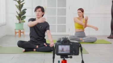 Genç bir kadın ve Asyalı erkek yoga yapıyor. Paspasın üzerinde el kası var. Evde kamerayla canlı yayın yapıyorlar..