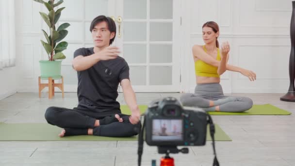 若い女性とアジアの男はマットの上にストレッチ手の筋肉でヨガを訓練し 自宅でカメラでライブストリーミング フィットネスでスポーツと活動を行うカップル 健康のための運動 — ストック動画