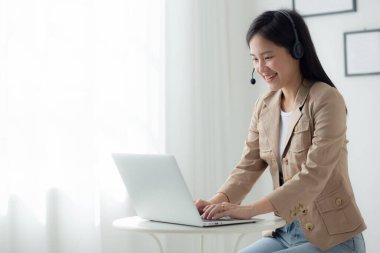 Young asian businesswoman wearing headset video call and explain conference online at home, woman wearing headphone meeting and talking for working from home, business and communication concept.