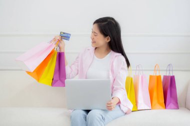 Kanepede oturan Asyalı genç bir kadın bilgisayarla online alışveriş yapıyor. Kredi kartıyla ve kesekağıdıyla ödeme yapıyor. İşletme konseptiyle ödeme yapan mutlu bir kadın..