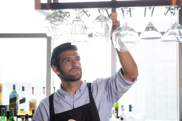 Young caucasian man is bartender holding and looking clean of glass with professional at counter, portrait handsome male preparation glass or wineglass, barman standing with confident at restaurant.