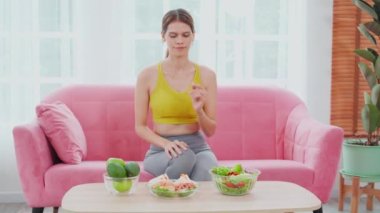 Güzel, genç, beyaz bir kadın kanepesinde oturmuş elinde kase sandviç ve sebze salatası tutarak evde yemek, sağlıklı beslenme, kilo kaybı ve yaşam tarzı kavramları için diyet yapıyor..