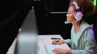 Kulaklık takan genç Afrikalı Amerikalı kadın video oyunu oynuyor ve evinde mikrofonla canlı yayın yapıyor, yarışma veya oyuncu seçimi, yayın ve eğlence konsepti için oyun ve esport oynuyor..