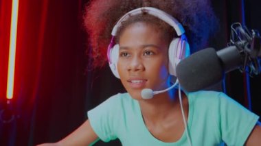 Kulaklık takan genç Afrikalı Amerikalı kadın video oyunu oynuyor ve evinde mikrofonla canlı yayın yapıyor, yarışma veya oyuncu seçimi, yayın ve eğlence konsepti için oyun ve esport oynuyor..