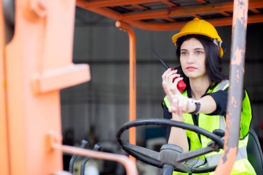 Young asian woman is foreman using radio for communication while driving forklift at distribution warehouse, engineer working and shipping container at factory, cargo and logistic, industry concept.