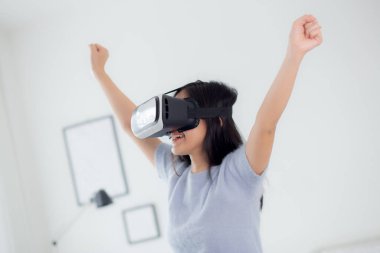 Young asian woman wearing vr headset playing game with excited in the bedroom at home, female using virtual reality or metaverse innovation for simulation 3D, lifestyles and technology concept.