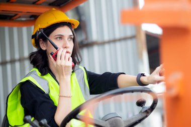 Young asian woman is foreman using radio for communication while driving forklift at distribution warehouse, engineer working and shipping container at factory, cargo and logistic, industry concept.