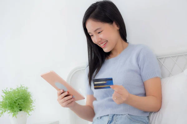 Young asian woman sitting on bed using digital tablet shopping online with credit card in the bedroom at home, female paying with transaction financial, purchase and payment, business concept.