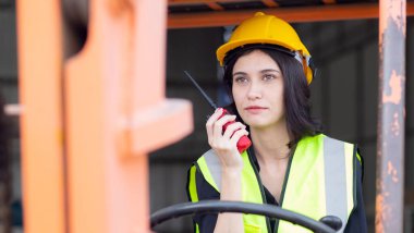 Young asian woman is foreman using radio for communication while driving forklift at distribution warehouse, engineer working and shipping container at factory, cargo and logistic, industry concept.