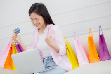 Kanepede oturan Asyalı genç bir kadın bilgisayarla online alışveriş yapıyor. Kredi kartıyla ve kesekağıdıyla ödeme yapıyor. İşletme konseptiyle ödeme yapan mutlu bir kadın..