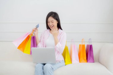 Kanepede oturan Asyalı genç bir kadın bilgisayarla online alışveriş yapıyor. Kredi kartıyla ve kesekağıdıyla ödeme yapıyor. İşletme konseptiyle ödeme yapan mutlu bir kadın..