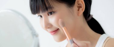 Güzel, genç Asyalı bir kadın. Yanak makyajına makyaj yapıyor. Yatakta kozmetik aynası, cilt bakımı ve yatak odasında kırışıklıklar olan güzel bir kız..