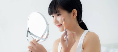 Güzel, genç Asyalı bir kadın. Yanak makyajına makyaj yapıyor. Yatakta kozmetik aynası, cilt bakımı ve yatak odasında kırışıklıklar olan güzel bir kız..