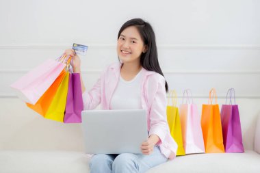 Kanepede oturan Asyalı genç bir kadın bilgisayarla online alışveriş yapıyor. Kredi kartıyla ve kesekağıdıyla ödeme yapıyor. İşletme konseptiyle ödeme yapan mutlu bir kadın..