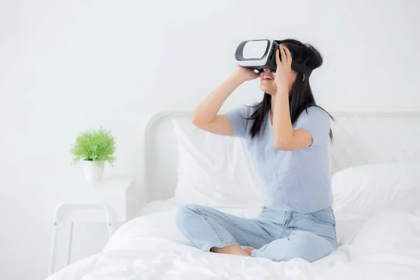 Stock image Young asian woman wearing vr headset playing game with excited on bed in the bedroom at home, female using virtual reality or metaverse innovation for simulation 3D, lifestyles and technology concept.