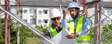 Two young architect discussion or contractor man while looking blueprint building structure together for survey on construction site, engineer planning project, industry and construction concept.