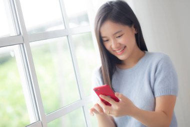 Genç Asyalı mutlu bir kadın evdeki oturma odasında akıllı telefonuyla sosyal ağa bakıyor, kadın gülümsüyor ve sosyal medyayı izliyor evde akıllı telefonuyla, yaşam tarzı konseptiyle.