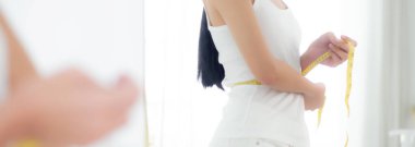 Güzel, genç Asyalı bir kadın ayakta duruyor ve ölçü alıyor. Kilo vermek için beliyle. Odada bir ayna var..