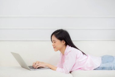 Genç Asyalı iş kadını evde dizüstü bilgisayarla çalışıyor oturma odasındaki kanepede internetten internete bağlanıyor. Kadın, rahat ve rahat bir şekilde kanepede uzanan not defterini kullanarak serbest çalışıyor..