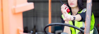 Young asian woman is foreman using radio for communication while driving forklift at distribution warehouse, engineer working and shipping container at factory, cargo and logistic, industry concept.