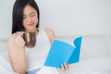 Young asian woman reading a book and holding coffee glass with relax and cozy on the bed in the bedroom at home, female leisure reading novel while resting, one person, lifestyles concept.