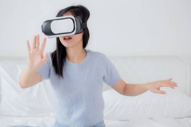 VR kulaklık takan genç Asyalı bir kadın evdeki yatak odasındaki yatakta heyecanla oyun oynuyor. Sanal gerçeklik ya da metaz yeniliklerini kullanarak 3 boyutlu, yaşam tarzları ve teknoloji kavramını kullanıyor..