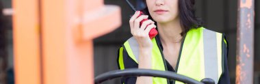 Young asian woman is foreman using radio for communication while driving forklift at distribution warehouse, engineer working and shipping container at factory, cargo and logistic, industry concept.