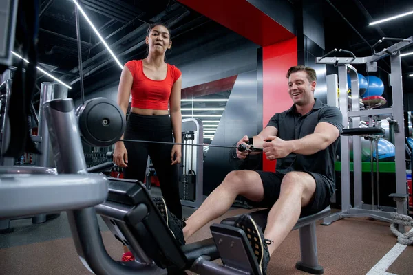Coach woman guiding workout with hands muscles stretch with equipment machine with man in fitness gym club, strength and flexibility training, man exercise with trainer or partnership, sport concept.