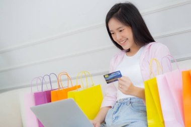 Kanepede oturan Asyalı genç bir kadın internet üzerinden dizüstü bilgisayar alışverişi yapıyor. İnternete kredi kartı alıyor, alışveriş, alım-satım ve ödeme ile ödeme yapan mutlu bir kız..