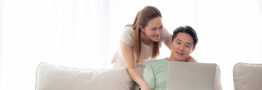 Genç Asyalı çift, sosyal medyayı veya videoyu izlemek için dizüstü bilgisayar kullanıyor. Evdeki oturma odasındaki kanepede rahatça oturuyor, kadın ve erkek bilgisayardan internete dizüstü bilgisayar izliyor, yaşam tarzı kavramı....