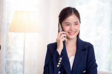 Güzel Asyalı genç danışman ya da iş kadını akıllı telefondan konuşuyor ve ev ofisinin oturma odasında dinlenirken gülümsüyor. Mutluluk acentesi ya da iş kadını akıllı telefondan konuşuyor..