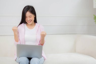 Happiness young asian woman sitting on sofa and working on laptop computer while successful and excitement at home, freelance female using notebook and achievement, business and lifestyles concept.