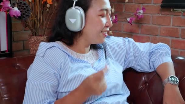 音楽を聴くためのヘッドフォンを身に着けている若いアジアの女性ハンディキャッパーは 自宅のリビングルームでソファーに座ってリラックスし 居心地の良いライフスタイルのコンセプトとソファに座って — ストック動画