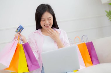 Kanepede oturan Asyalı genç bir kadın bilgisayarla online alışveriş yapıyor. Kredi kartıyla ve kesekağıdıyla ödeme yapıyor. İşletme konseptiyle ödeme yapan mutlu bir kadın..