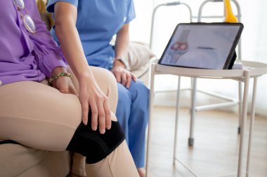 Evde tablet oturma odası, bakıcı ya da fizyoterapist olan yaşlı kadını açıklarken, tendon ve dizdeki artrit ve bacağındaki teşhis ve rehabilitasyonu kontrol etmeye yardım eden bakıcı ya da hemşire.