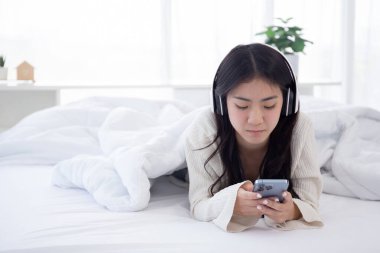 Güzel Asyalı genç bir kadın müzik için kulaklık takıyor. Yatak odasında yatakta dinlenirken akıllı telefon kullanıyor. Kulaklık takıyor ve hafta sonları rahat bir şekilde akıllı telefon izliyor..