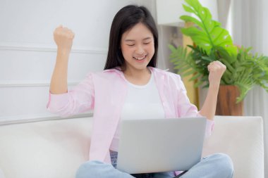 Happiness young asian woman sitting on sofa and working on laptop computer while successful and excitement at home, freelance female using notebook and achievement, business and lifestyles concept.