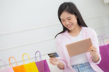 Young asian woman sitting on sofa using digital tablet shopping online with credit card while paper bag on couch, female paying with transaction financial, purchase and payment, business concept.